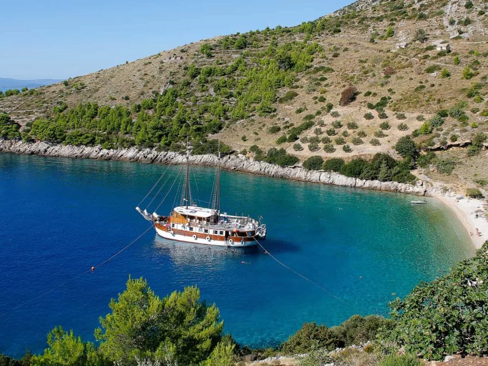 Skrivena uvala na otoku Braču, Hrvatska, s tirkiznim morem, šljunčanom plažom i usidrenim drvenim jedrenjakom. Okružena je zelenim brežuljcima prekrivenima borovima i mediteranskim raslinjem.