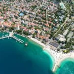 Zračni pogled na Crikvenicu, Hrvatska, s lukom punom brodova, šljunčanim plažama i kristalno čistim morem. Naselje s crvenim krovovima prostire se duž obale, okruženo zelenilom i turističkim sadržajima.