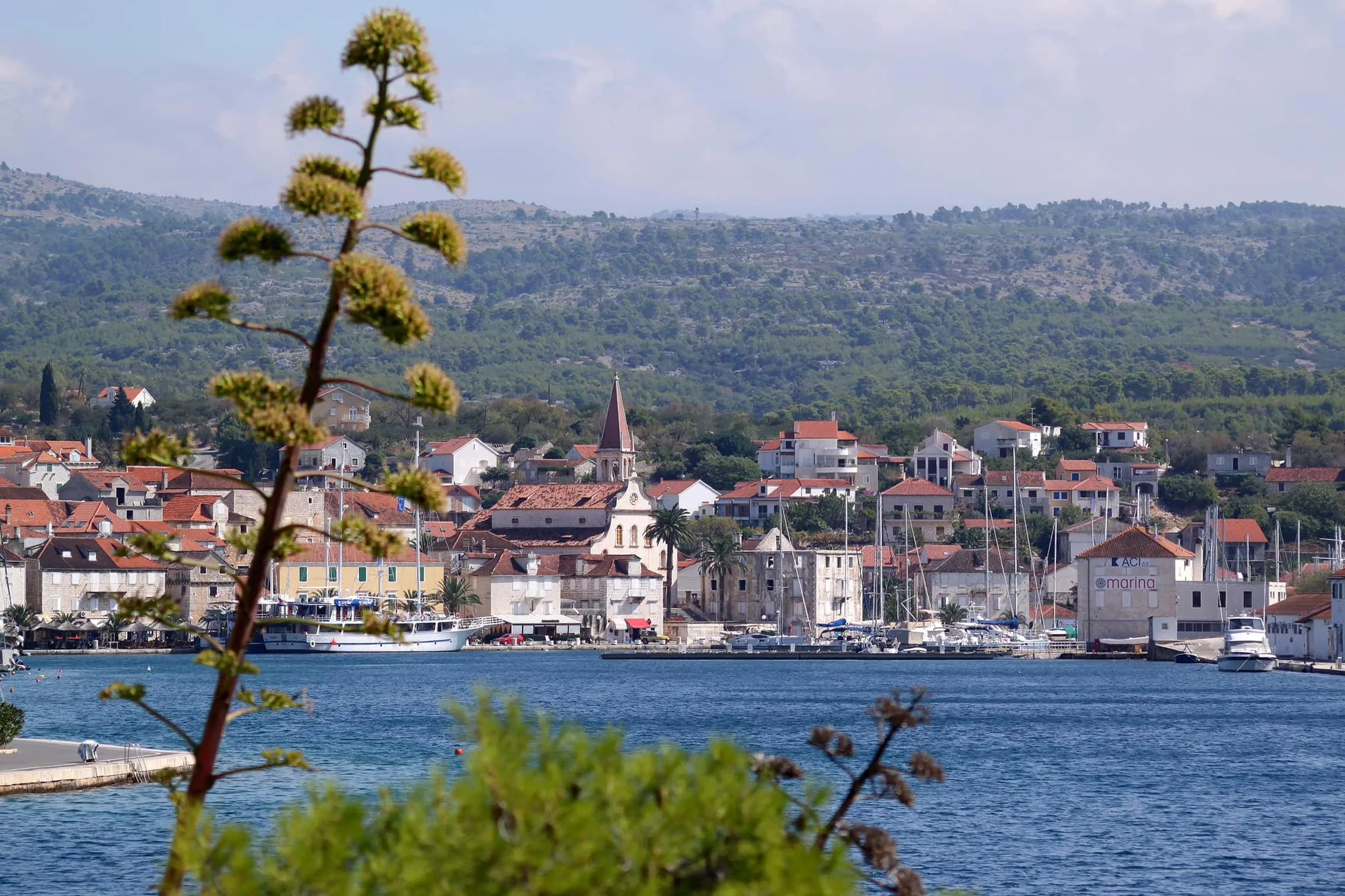 Kilátás Milna kikötőjére, Brač szigetén, Horvátország, a Gospe od Blagovijesti templommal, kőházakkal és jachtokkal teli marinával. Az előtérben egy agávé növény emelkedik ki, míg a háttérben zöld dombok láthatók.