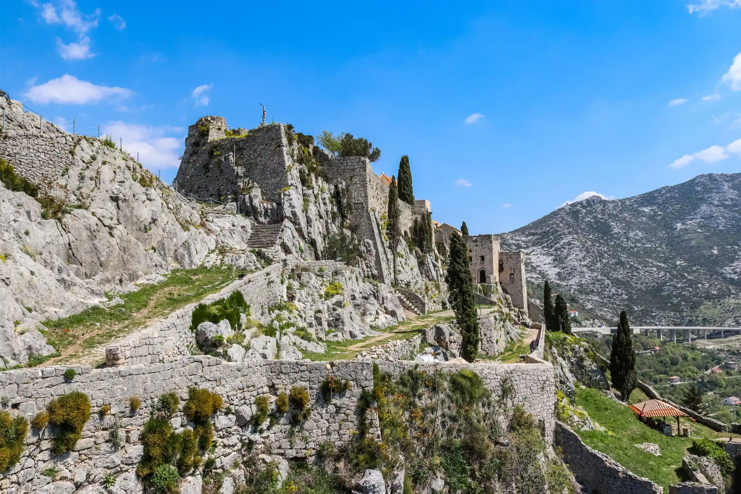 Pevnosť Klis na strmom brale s kamennými hradbami, úzkymi chodníkmi a cyprusmi, s horami v pozadí