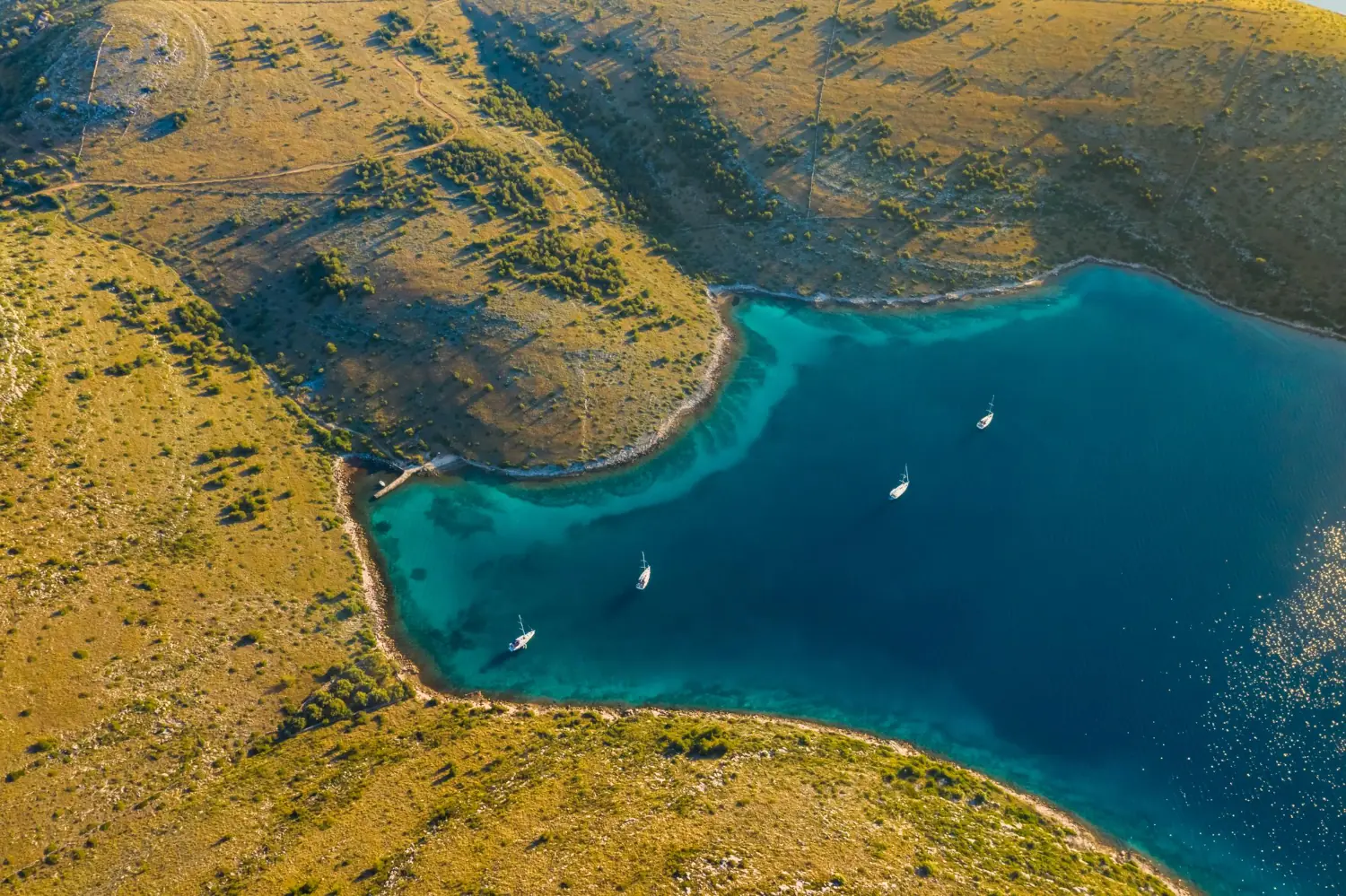 Aerial view of a turquoise bay in the Kornati with moored sailboats and dry-stone walls on karst terrain.