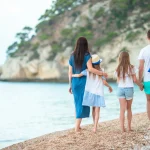 Familienurlaub in Croatia – eine vierköpfige Familie spaziert an einem ruhigen Kiesstrand am Meer entlang, vor einer felsigen Küste in Kroatien.