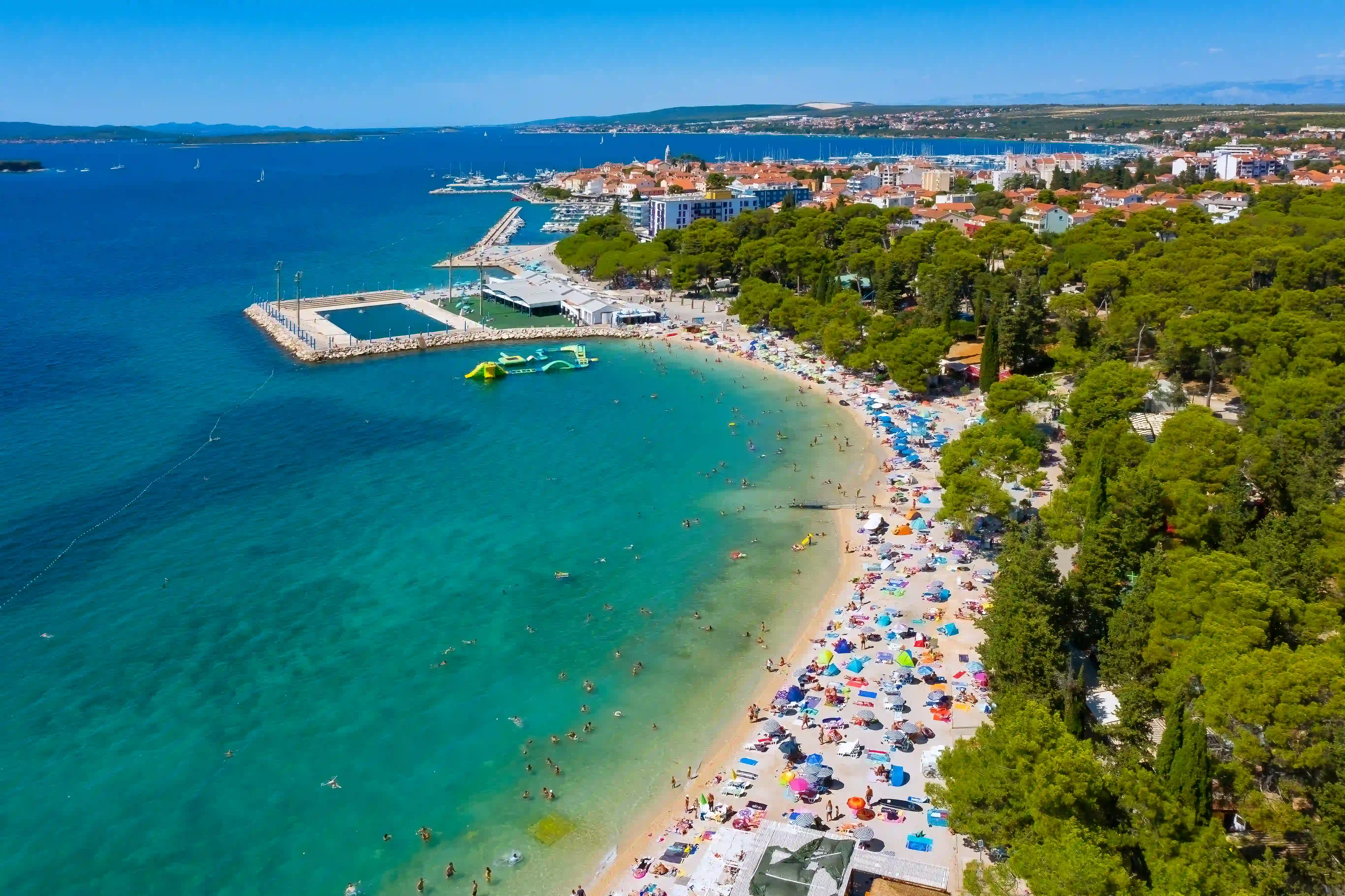 Panoramski pogled na plažo Dražica v Biogradu na Moru s kopalci in turkiznim morjem.