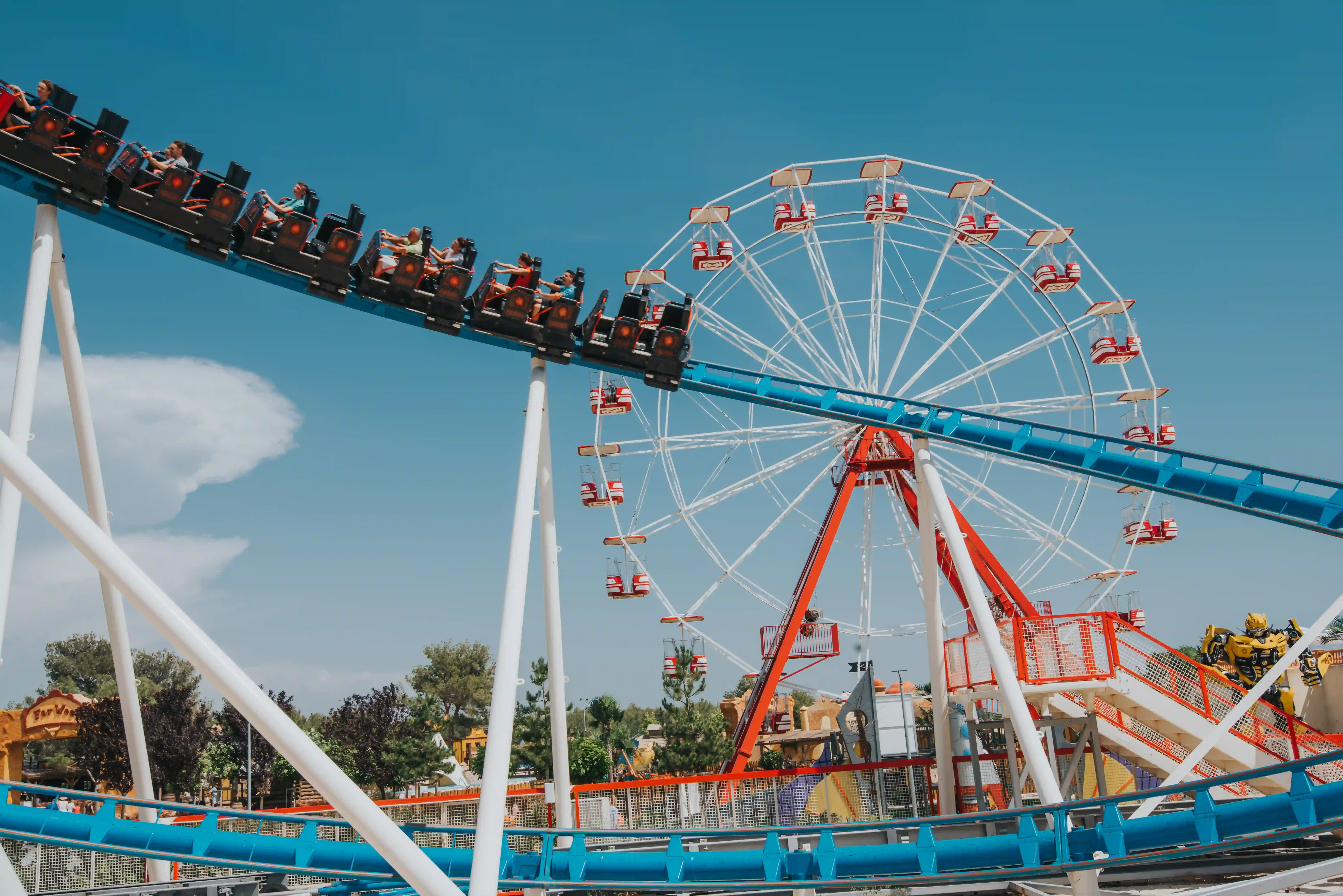 Vlak smrti v vožnji pred velikim belim panoramskim kolesom v zabaviščnem parku Dalmaland pod jasnim nebom.