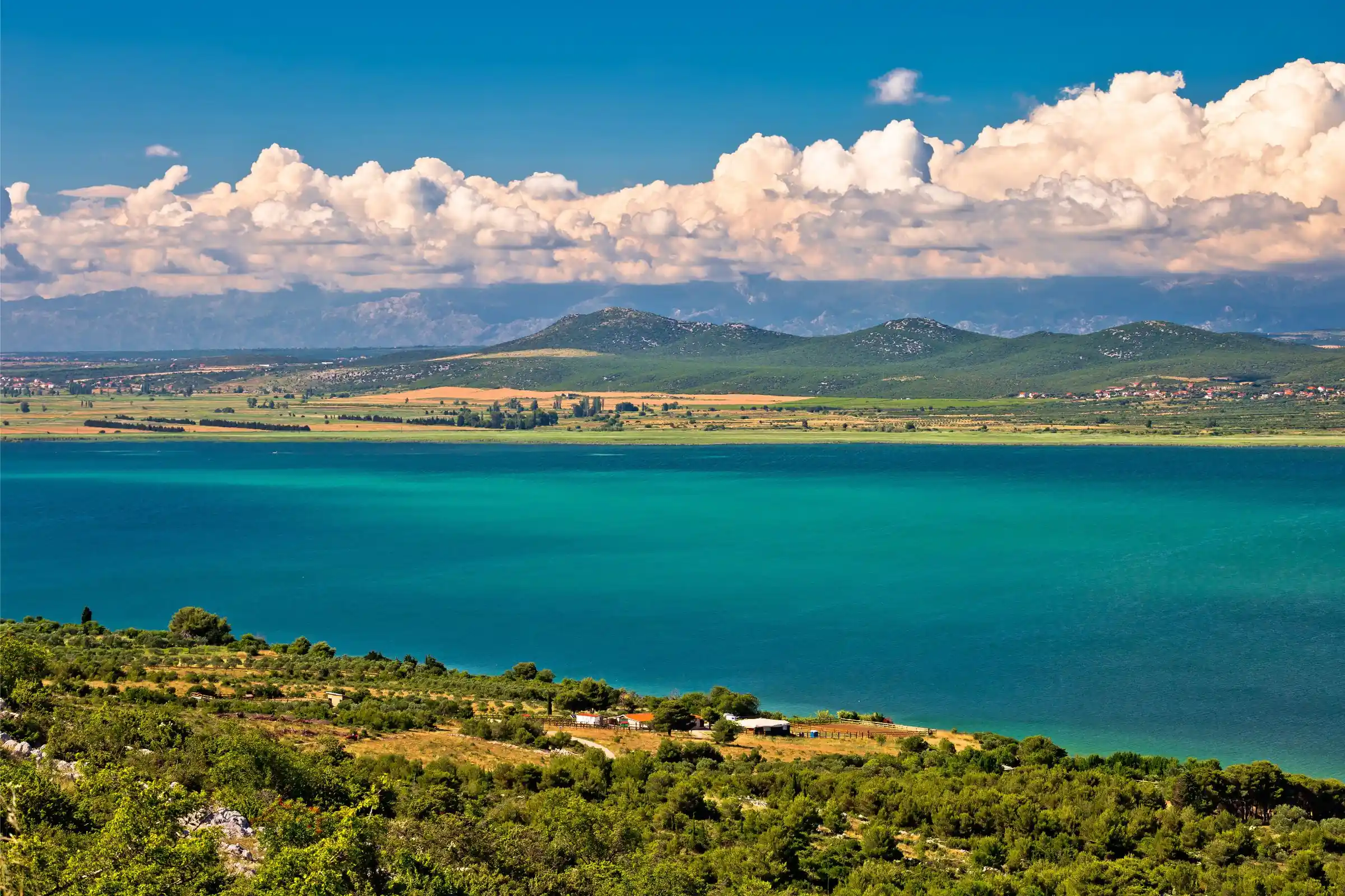 Panoramski pogled na Park narave Vransko jezero s turkizno vodo, zelenimi griči in belimi oblaki v ozadju.
